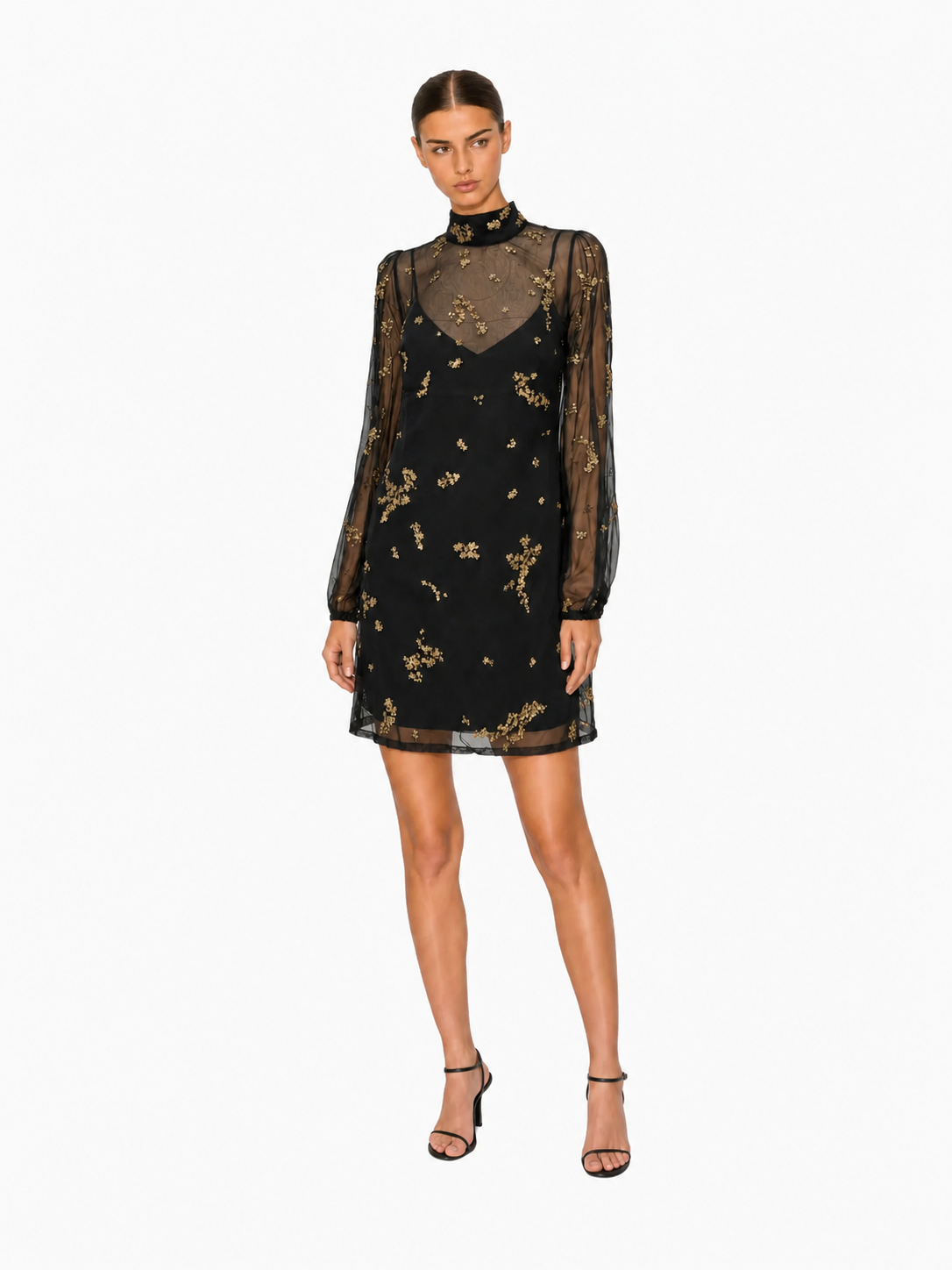 A model wearing a black mini dress with sheer long sleeves, a high neck, and gold floral embroidery, paired with black strappy heels, on a light gray background.